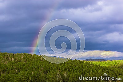 Part of colored rainbow on dark thunder clouds with green forest mountain background. horizontal view. Stock Photo
