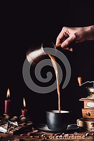 partial image of woman pouring coffee from turk into cup at table with chocolate, truffles, candles and coffee grains Stock Photo
