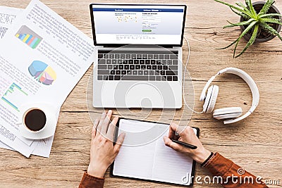 partial view of man making notes in notebook at workplace with laptop with facebook logo, papers, cup of coffee Editorial Stock Photo