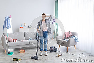 After party chaos. Dismayed millennial woman standing in her messy apartment with vacuum cleaner, holding head in terror Stock Photo