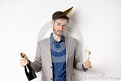 Party guy standing in birthday hat and celebrating, holding champagne bottle and glass, looking drunk, wearing grey suit Stock Photo