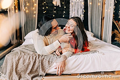 Passionate romantic couple spending home with garlands. Young woman and man are lying on bed in sweaters and hugging Stock Photo