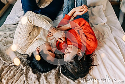 Passionate romantic couple spending home with garlands. Young woman and man are lying on bed in sweaters and hugging Stock Photo