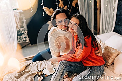 Passionate romantic couple spending home with garlands. Young woman and man are lying on bed in sweaters and hugging Stock Photo