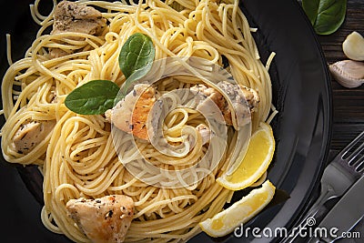 Pasta , spaghetti with chicken , lemon slices, garlic, greens, spinach, fork, on a black plate on dark , close up Stock Photo