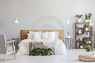 Patterned armchair near white wooden bed in grey bedroom interior with pouf and plants. Real photo Stock Photo