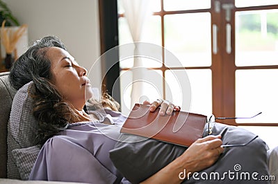 Peaceful and relaxed aged Asian woman fall asleep while reading a book on her comfortable sofa Stock Photo
