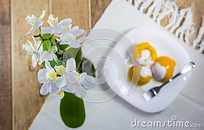 Peach in syrup with cream milk, dessert served on a white dish on a table decorated with vase of orchids Stock Photo