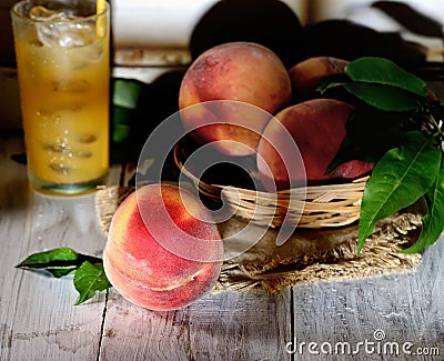Peaches on a branch with leaves in a wicker from rods bowl and a glass of peach juice on a light wooden background Stock Photo