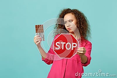 Pensive african girl in pink casual clothes holding chocolate bar, text board STOP isolated on blue turquoise wall Stock Photo