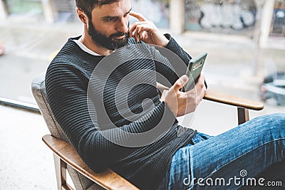 Pensive bearded man sitting home and using mobile smartphone. Man using gadget at hotel lobby. Horizontal. Blurred Stock Photo