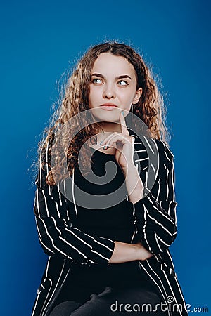 pensive curly girl looks up and thinks about the future on a blue background in a striped jacket Stock Photo