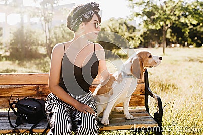 Pensive lady wears black ribbon in hair looking away while sitting on wooden bench with beagle dog. Outdoor portrait of Stock Photo