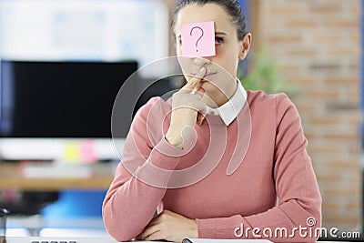 Pensive young woman sitting at table with sticker with question mark on her forehead Stock Photo