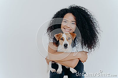 People, animal or pet care concept. Curly haired woman embraces favourite dog, smiles pleasantly, stands against white background Stock Photo