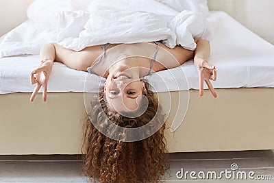 People, bedding, fun concept. Lovely delighted female with curly hair, keeps head down, makes peace gesture, lies on comfortable b Stock Photo