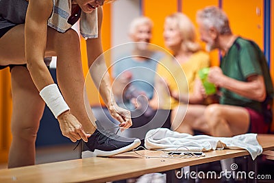 People in gym locker room. Young woman tying her shoe, seniors in the backgorund sitting and talking Stock Photo