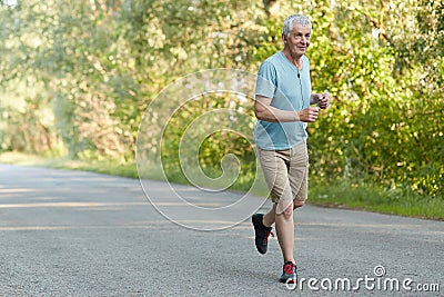 People and healthy lifestyle concept. Handsome wrinkled mature male joggs quickly on road, listens pleasant music in earphones, br Stock Photo