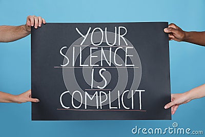 People holding sign with phrase Your Silence Is Complicit on light blue background, closeup. Racism concept Stock Photo