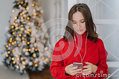 People and holidays concept. Concentrated dark haired young woman holds cell phone in hands, reads news in internet, uses earphone Stock Photo