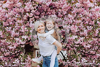 People, holidays and family concept - daughter hugs mom sitting on her back on cherry blossom background. Walk in the sakura trees Stock Photo