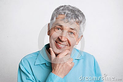 People, pension and positive emotions concept. Pleased mature male with grey hair, keeps hand under chin, wears formal blue shirt, Stock Photo