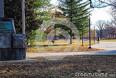 People sitting in a swing in the park surrounded by lush green trees and plants and autumn colored trees and other people walking Editorial Stock Photo