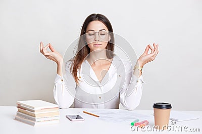 People and spirituality concept. Relaxed brunette young woman poses at workplace in mudra sign, enjoys peaceful atmosphere, pull h Stock Photo