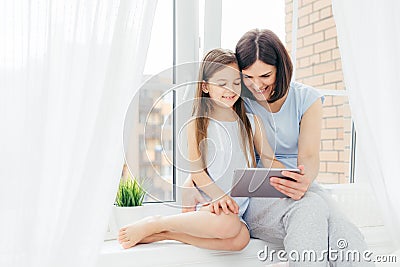 People, technology, family, children concept. Positive young other and her small daughter sit on window sill, hold digital tablet, Stock Photo