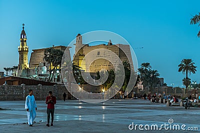 People walking in the square in front of the ancient illuminated Luxor temple at night with dark blue sky and glowing brickwalls Editorial Stock Photo