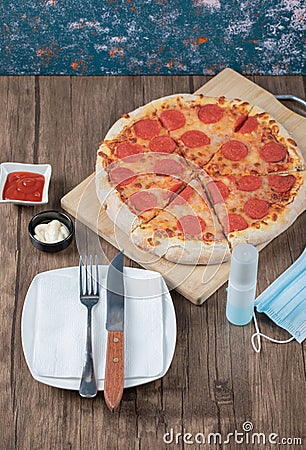 Pepperoni pizza on a wooden board with sauces, plate, hand sanitizer Stock Photo