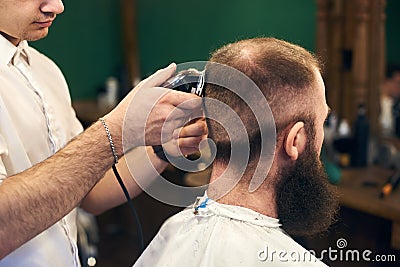 Professional male short haircutting for bearded client in barbershop. Young barber working with electric razor. Close up Stock Photo