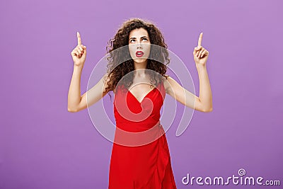 Perplexed focused attractive european woman with curly hairstyle in red evening dress open mouth raising arms looking Stock Photo