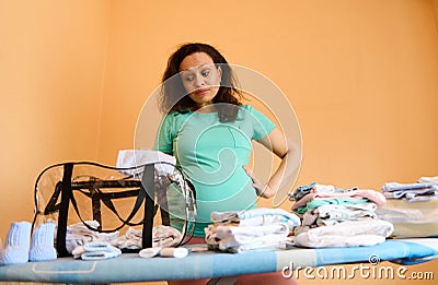 Perplexed young pregnant woman, expecting baby, collecting newborn clothes in the bag for maternity hospital Stock Photo