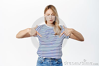 Perplexed young woman grimacing, pointing and looking down with skeptical, doubtful face, dislike smth, feeling Stock Photo