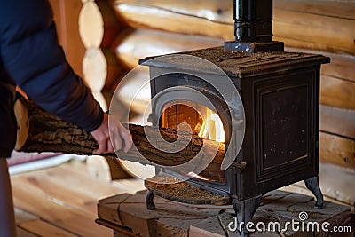 person feeding a log into a castiron stove in a log cabin interior Stock Photo