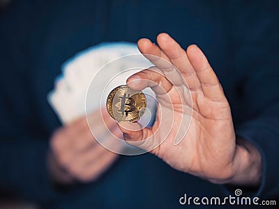 Person holding bitcoin in one hand, pile of cash money in other hand. Invest into virtual currency concept. Selective focus. Dark Stock Photo