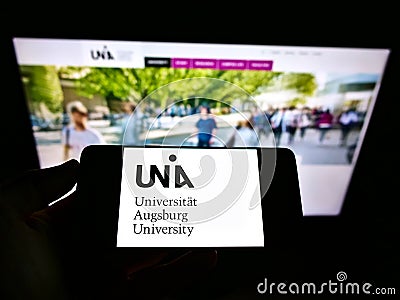 Person holding cellphone with logo of German university UniversitÃ¤t Augsburg on screen in front of webpage. Editorial Stock Photo