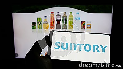 Person holding cellphone with logo of Japanese beverage company Suntory Holdings K.K. on screen in front of webpage. Editorial Stock Photo