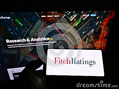 Person holding mobile phone with logo of American credit rating agency Fitch Ratings Inc. on screen in front of web page. Editorial Stock Photo