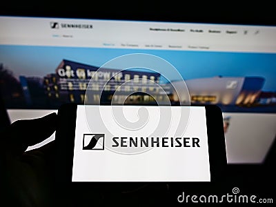 Person holding mobile phone with logo of German audio company Sennheiser electronic GmbH Co KG on screen in front of webpage. Editorial Stock Photo