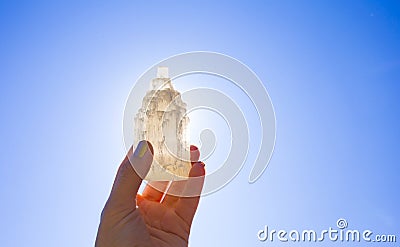 Person holding naturally carved mineral stone Selenite tower against sun and blue sky, stone has healing and cleansing properties. Stock Photo