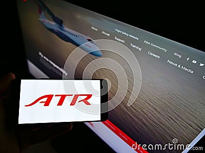 Person holding phone with logo of aircraft manufacturer Avions de Transport RÃ©gional (ATR) on screen with web page. Editorial Stock Photo