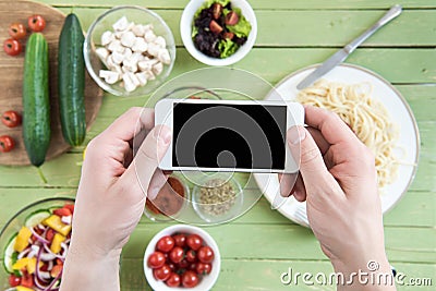 Person holding smartphone with blank screen and photographing spaghetti and fresh vegetables on wooden table Stock Photo