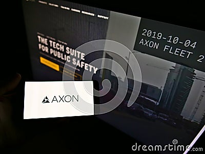 Person holding smartphone with logo of American weapon manufacturer Axon Enterprise Inc. on screen in front of website. Editorial Stock Photo