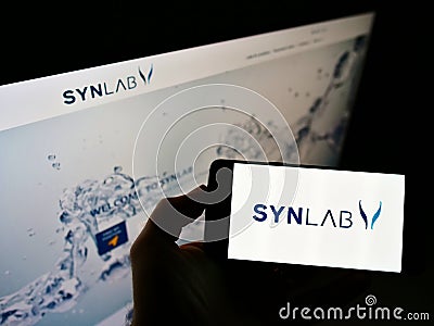 Person holding smartphone with logo of German medical diagnostics company Synlab Group on screen in front of website. Editorial Stock Photo