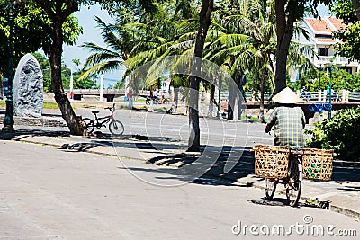 Person seen from his back moving on a bicycle wearing a conical hat and a carrying pole with vegetables. Empty street. Hue, Editorial Stock Photo