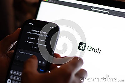 Tech guy trying out Grok in mobile phone with the laptop screen showing Grok logo, an generative, conversational, neural network, Editorial Stock Photo