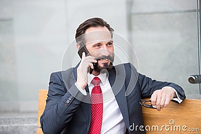 On the phone. Portrait of modern businessman talking on smart-phone while sitting on bench outdoors. bearded man holding glasses Stock Photo