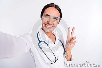 Photo of adorable cheerful young woman doctor wear formal coat tacking selfie smiling showing v-sign isolated white Stock Photo
